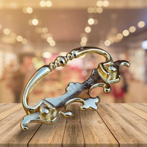 May include: An ornate metal drawer pull in a gold tone, featuring a decorative design. The pull has a curved handle and intricate details.