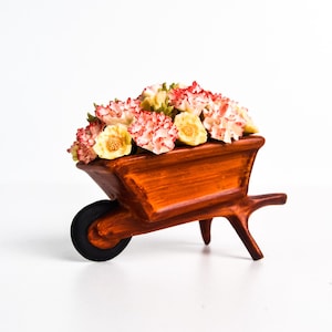 May include: A small, wooden wheelbarrow ornament filled with flowers. The wheelbarrow is painted a warm brown colour and has a single black wheel. The flowers are a mix of red, white, and yellow, creating a colourful display.
