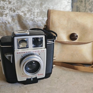 May include: A vintage Kodak Brownie Twin 20 camera, black and silver, with a matching tan leather case. The camera features the text "Brownie Twin 20" and "Kodak". The case has a brown button closure and a strap.