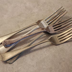 May include: Several silver forks with tines and long handles are arranged in a slightly overlapping pattern. The forks have a vintage appearance. The metal has a slightly aged appearance, suggesting vintage or antique pieces.