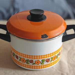May include: A vintage orange and white enamel pot with a lid and two black handles. The lid is orange with a black knob. The pot has a white body with an orange floral and checkered pattern around the base. A charming kitchenware item.