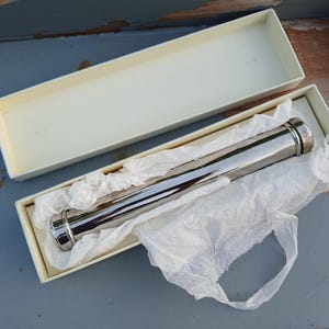 May include: A cylindrical, polished silver-tone metal tube rests inside an off-white rectangular cardboard box. The tube is partially wrapped in crumpled white tissue paper, with the lid of the box placed separately above the base container.