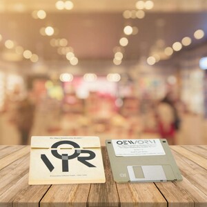 May include: Two vintage computer items on a wooden surface. One is a beige square with the text "VOR" in black, and the other is a 8.89 cm floppy disk with a label. The background is blurred.