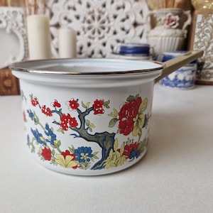 May include: White enamel saucepan with a floral design featuring red, yellow, blue, and green flowers. The saucepan has a silver rim and a beige handle.