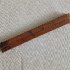 May include: A vintage wooden folding ruler, with a warm brown colour and brass-coloured metal accents. The ruler is folded, showing its segmented design. The wood has a slightly worn appearance, suggesting age and use. The ruler is approximately 15 cm long when folded.