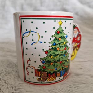 May include: White ceramic Christmas mug with a green, red, and yellow Christmas tree, a teddy bear, and a Santa Claus figure. The mug has a red, green, and white border.