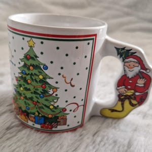 May include: White ceramic Christmas mug with a green Christmas tree and red and white presents on the front. The handle is shaped like a Santa Claus boot.