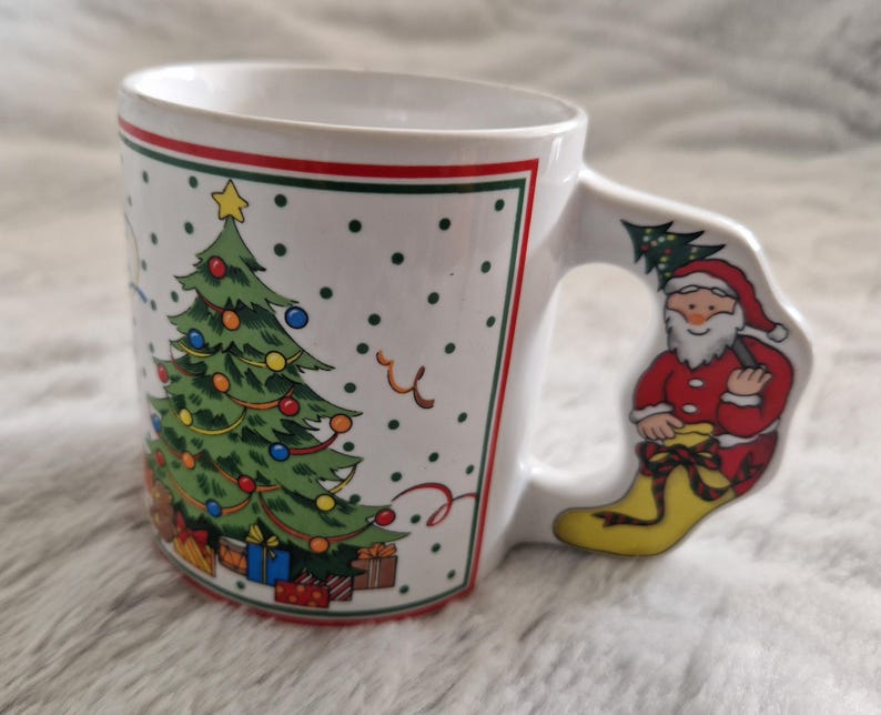 May include: White ceramic Christmas mug with a green Christmas tree and red and white presents on the front. The handle is shaped like a Santa Claus boot.