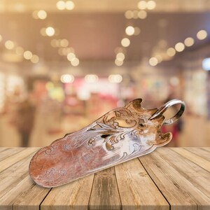 May include: An ornate silver serving utensil with a decorative handle and a tarnished finish. The utensil is lying on a wooden surface.