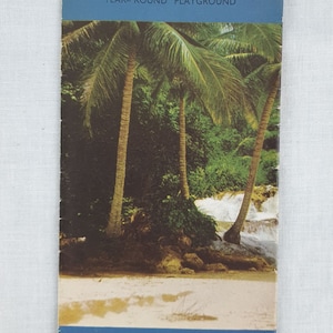 May include: A vintage travel brochure for Jamaica, featuring a photo of palm trees and a waterfall. The text on the brochure reads "Jamaica, Year-Round Playground."