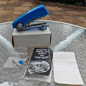May include: A blue handheld sewing machine with a silver needle and thread, on a white cardboard box. The machine has wheels and a small blue knob. Instructions for handling the machine are included.