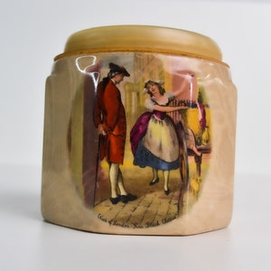 May include: A ceramic jar with a lid, featuring a colourful illustration of a man and woman in 18th-century clothing. The jar has a hexagonal shape and the text "Alice of London - Fine Black China" is printed on the bottom.