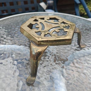 May include: A vintage brass trivet with a hexagonal top and three curved legs. The trivet features an openwork design on the top surface. The brass has a warm, aged patina. This decorative item is designed to protect surfaces from hot cookware.