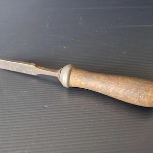 May include: A vintage wood chisel with a metal blade and a wooden handle. The chisel has a slightly curved handle and a metal ferrule where the blade meets the handle. The blade is angled and appears to be well-worn.