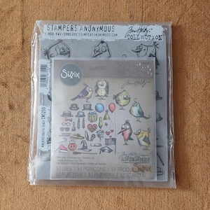 May include: A Stampers Anonymous Jim Holtz collection clear stamp set and a Sizzix Framelits die set featuring 51 metal dies. The packaging displays various illustrated birds, hats, glasses, ties, and party accessories in a whimsical sketch style.