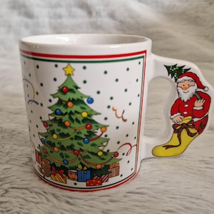 May include: White ceramic Christmas mug with a green Christmas tree and red and white Santa Claus on the handle. The mug has a red and green trim around the top.