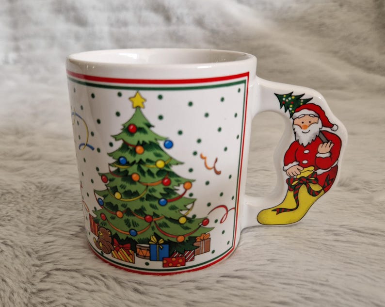 May include: White ceramic Christmas mug with a green Christmas tree and red and white Santa Claus on the handle. The mug has a red and green trim around the top.