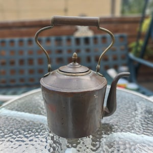 May include: A copper kettle with a brass handle and spout. The kettle has a rounded body and a lid with a small finial. The handle is made of wood. The kettle is likely used for boiling water.