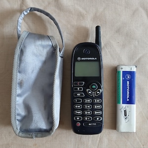 May include: A dark blue Motorola m3788 mobile phone with an external antenna, a silver fabric carrying case with a handle, and a white and blue 4.8V Nickel-Metal Hydride battery pack with Motorola branding and a diagram for installation.