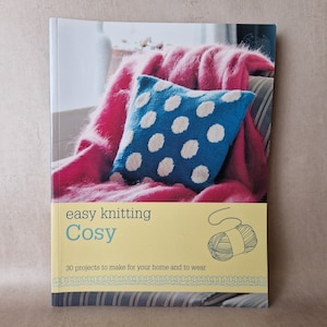 May include: A book titled "easy knitting Cosy" with a yellow background and the text "30 projects to make for your home and to wear". The cover features a pink fluffy throw blanket and a blue knitted cushion with white polka dots.