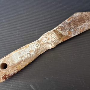 May include: A worn wooden tool with a flat, blade-like end and a handle with a hole. The tool has a rustic appearance with signs of age and use. The color is a mix of brown, beige, and white.
