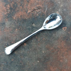 May include: A small, shiny silver spoon with a rounded bowl and a long, slender handle. The spoon reflects the surrounding environment, creating a mirror-like effect. The spoon is likely made of stainless steel.