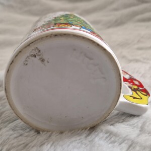 May include: White ceramic Christmas mug with a colorful illustration of a Christmas tree and figures. The mug has a handle shaped like a Santa Claus figure.