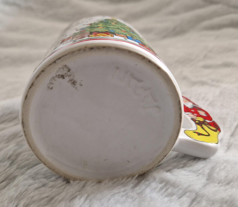 May include: White ceramic Christmas mug with a colorful illustration of a Christmas tree and figures. The mug has a handle shaped like a Santa Claus figure.