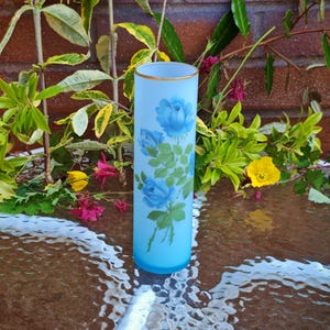 May include: A light blue glass vase with a gold rim and a floral design of blue roses and green leaves. The vase is cylindrical and stands upright, showcasing a classic floral pattern. The background includes greenery and a brick wall.