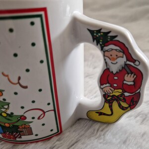 May include: White ceramic Christmas mug with a red and green border. The mug features a Christmas tree with presents and a Santa Claus holding a tree on the handle.