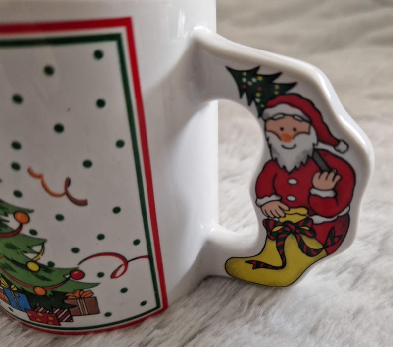 May include: White ceramic Christmas mug with a red and green border. The mug features a Christmas tree with presents and a Santa Claus holding a tree on the handle.