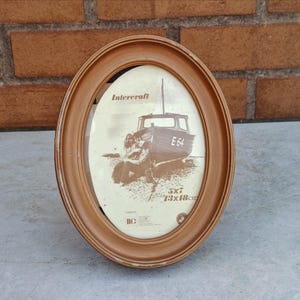 May include: Oval light brown picture frame with a distressed finish. The frame holds a sepia-toned print of a boat with the text "E64". The frame is 13x18 cm. The brand name "Intercraft" is visible. The frame has a vintage aesthetic.