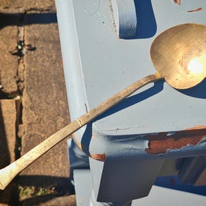 May include: A vintage brass ladle with a long handle and a rounded bowl. The ladle has a golden hue and shows signs of age and use. The handle has a textured design. The ladle is resting on a weathered, light blue surface.