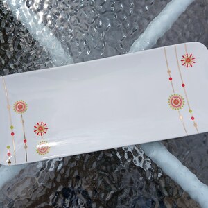 May include: A white rectangular serving platter with rounded corners. It features a floral design in red, gold, and green. The design includes stylized flowers and dotted lines. The platter is likely ceramic. Measurements are approximately 30 cm long.