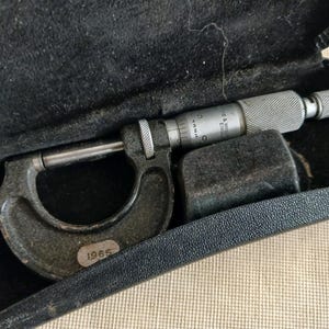 May include: A vintage micrometer in a black case. The micrometer is made of metal and has a silver finish with a knurled grip. The case is lined with black fabric. The micrometer is marked with the year 1965.