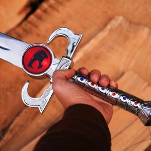 May include: A silver sword with a polished blade and ornate handle. The handle features red and blue accents. A red circular emblem with a black cat-like figure is on the blade. The sword is held against a wooden background.