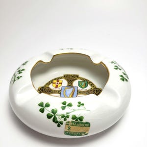May include: White ceramic ashtray with a gold rim and green shamrock designs. The center features a decorative emblem with a harp and shields. The ashtray has the text "HANNON FREEPORT" on a sticker.