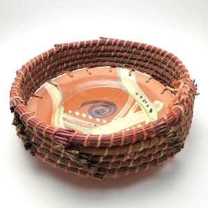 May include: A handcrafted, round basket with a painted ceramic base. The basket is woven with reddish-brown and tan natural fibers. The ceramic base is orange with abstract blue, white, and green painted designs. The basket is approximately 6 inches in diameter.