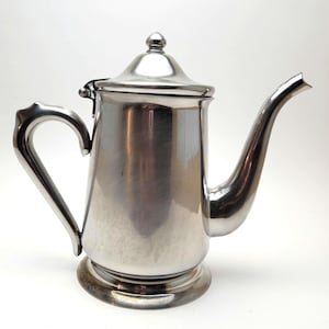 May include: A polished silver coffee pot with a curved spout and a handle. The pot has a lid with a small knob on top and sits on a round base. The design is classic and elegant, suitable for serving coffee or tea.