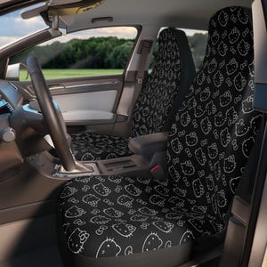 Pode incluir: Capas de assento de carro pretas com um padrão de contorno branco repetido de um gato de desenho animado com um laço. As capas são instaladas no interior de um carro, com o volante e o painel visíveis. O fundo mostra uma vista de um campo verde e árvores.