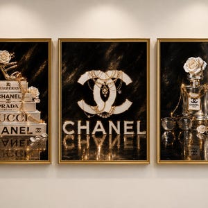 May include: A set of three framed wall art prints featuring luxury fashion themes. The first shows stacked books with a gold high-heeled shoe and white roses. The second displays a large Chanel logo with gold chains. The third shows a perfume bottle.