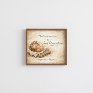 May include: A framed print showing a loaf of bread on a wooden board and a linen cloth. The print has text that reads "Slow hands, warm hearts, and bread that rises with time." The print is in a brown wooden frame.
