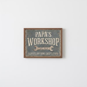 Puede incluir: Un letrero enmarcado con las palabras "Papa's Workshop" en un estilo vintage. El letrero incluye un gráfico de llave inglesa y el texto "Est. 2026". La parte inferior del letrero dice "Puedo arreglar cualquier cosa excepto la estupidez".