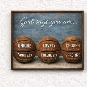 Puede incluir: Arte de pared enmarcado en madera con un fondo azul y el texto "Dios dice que eres...". Se presentan tres balones de baloncesto, cada uno con una palabra: UNIQUE, LOVELY y CHOSEN, junto con versículos bíblicos relacionados. El marco es marrón.