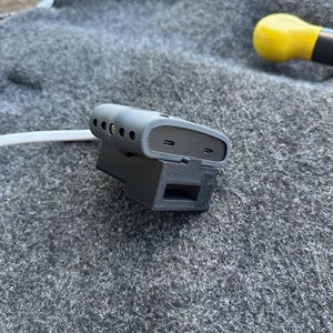 Puede incluir: Un dispositivo electrónico gris de forma rectangular redondeada con dos puertos USB-C en el lateral, montado en un soporte de plástico negro impreso en 3D. Un cable blanco sale del lateral de la unidad gris, que presenta una serie de pequeños orificios.