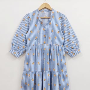 May include: A midi-length dress featuring a light blue and white vertical striped pattern with a repeating yellow lemon print. The dress has three-quarter length sleeves, a tiered skirt, and a V-neckline with a tie closure at the collar.