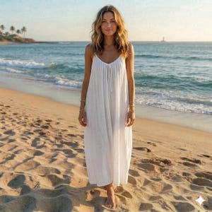 May include: A white, flowing sundress with spaghetti straps and a drawstring neckline. The dress is worn on a sandy beach with the ocean in the background. The dress appears to be made of a lightweight fabric.