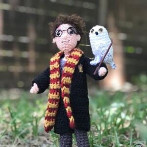 May include: A crocheted Harry Potter doll wearing a black robe and a red and gold scarf, holding a white owl with yellow eyes. The owl is holding a stick in its beak.