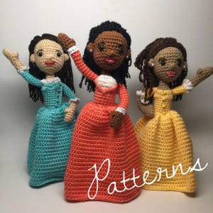 May include: Three crocheted dolls dressed in colonial-style dresses. The dolls are wearing teal, orange, and yellow dresses. The dolls are standing on a white surface. The word "Patterns" is written in white on a red background in the lower right corner of the image.