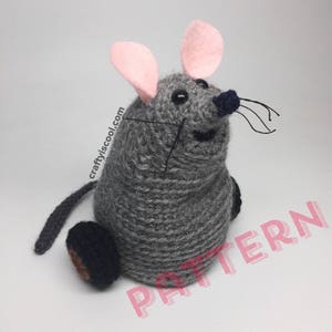 May include: A grey crocheted mouse with pink ears and black eyes. The mouse has a black nose and whiskers. The mouse is sitting on a white surface.
